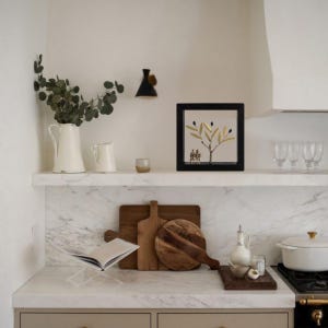 Op de afbeelding: Een wit marmeren aanrecht met een witte keramische kan, een witte keramische roomkan, een kleine kaars, een ingelijst kunstwerk van een gouden olijftak, twee houten snijplanken, een glazen fles met een kurk, een witte keramische ovenschaal en een zwart en zilveren fornuis.