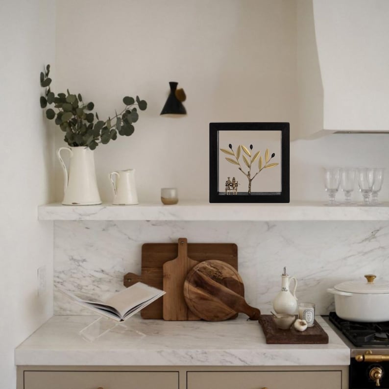 Op de afbeelding: Een wit marmeren aanrecht met een witte keramische kan, een witte keramische roomkan, een kleine kaars, een ingelijst kunstwerk van een gouden olijftak, twee houten snijplanken, een glazen fles met een kurk, een witte keramische ovenschaal en een zwart en zilveren fornuis.