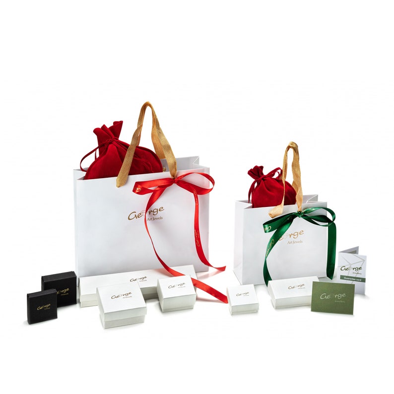 Op de afbeelding: Twee witte cadeauzakken met rode en groene strikken. De zakken bevatten rode fluwelen zakjes. De zakken staan op een wit oppervlak met verschillende kleine witte dozen en &eacute;&eacute;n zwarte doos. Op de zakken staat "George Art Jewels" gedrukt.