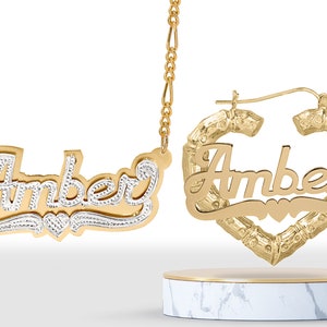 May include: Gold nameplate necklace and earrings with the name "Amber" in a cursive font. The necklace has a delicate chain and the earrings are hoop style with a heart-shaped design.