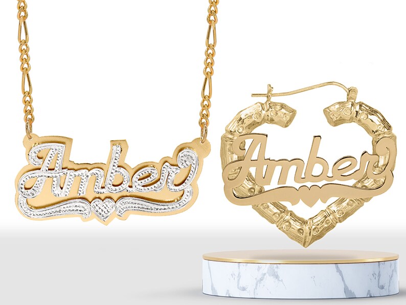 May include: Gold nameplate necklace and earrings with the name "Amber" in a cursive font. The necklace has a delicate chain and the earrings are hoop style with a heart-shaped design.