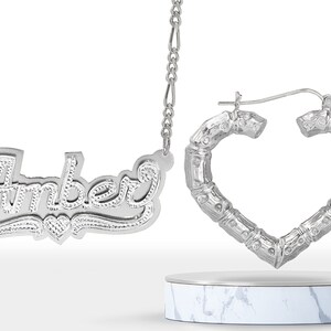 May include: Silver necklace with the name "Amber" spelled out in cursive script and a small heart charm.  The necklace is paired with silver heart-shaped hoop earrings with a bamboo-like design.