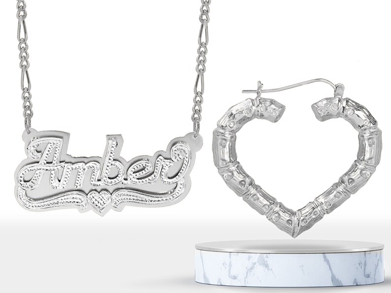 May include: Silver necklace with the name "Amber" spelled out in cursive script and a small heart charm.  The necklace is paired with silver heart-shaped hoop earrings with a bamboo-like design.