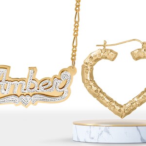 May include: Gold necklace with a nameplate pendant that says "Amber" in a cursive font with a small heart charm. The necklace is paired with gold heart-shaped hoop earrings.