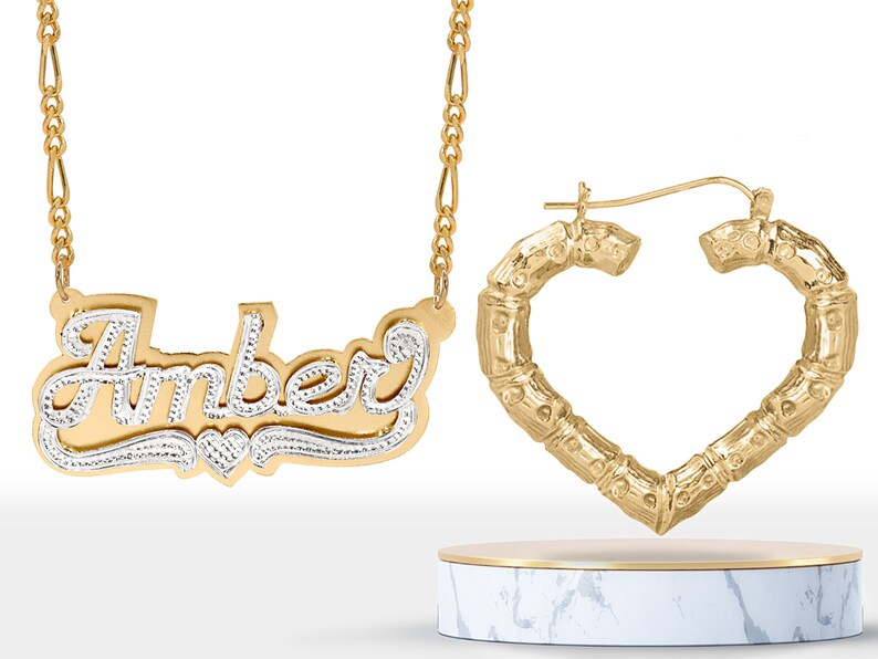 May include: Gold necklace with a nameplate pendant that says "Amber" in a cursive font with a small heart charm. The necklace is paired with gold heart-shaped hoop earrings.