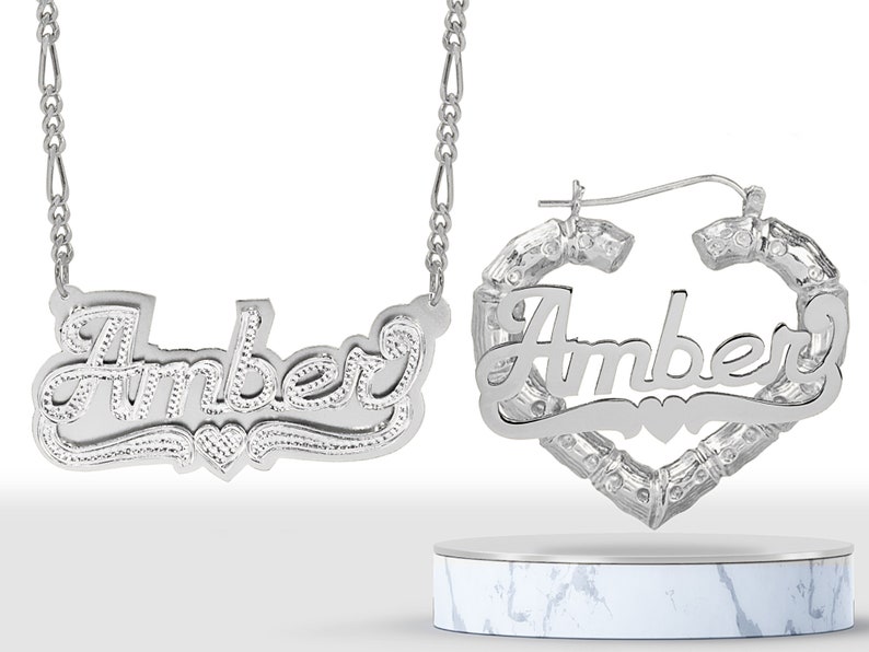 May include: Silver necklace and hoop earrings with the name "Amber" in a cursive font with a heart design.