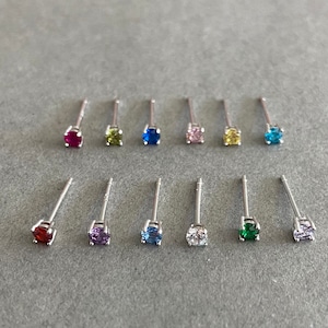 May include: A set of 12 silver stud earrings with colorful gemstones. The earrings are arranged in two rows of six, with each row featuring a different color gemstone. The colors include red, green, blue, pink, yellow, and clear.