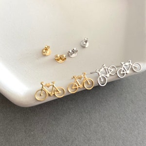 May include: Two pairs of bicycle-shaped stud earrings, one pair in a gold tone and one pair in a silver tone. Each earring features a small heart detail on the frame and includes a matching metal earring back for secure fastening.
