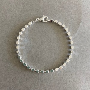 Puede incluir: Un brazalete de plata con un diseño de cadena de eslabones cuadrados. El brazalete tiene un cierre de langosta.