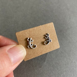 May include: A pair of silver-colored lizard stud earrings displayed on a small, brown cardboard tag. The earrings are detailed with textured scales and curled tails. The background is a neutral gray.