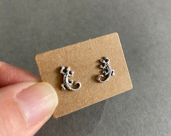May include: A pair of silver-colored lizard stud earrings displayed on a small, brown cardboard tag. The earrings are detailed with textured scales and curled tails. The background is a neutral gray.