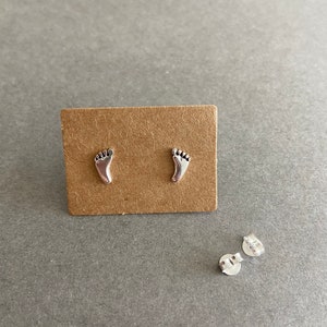 May include: A pair of silver stud earrings shaped like footprints. The earrings are displayed on a brown card.