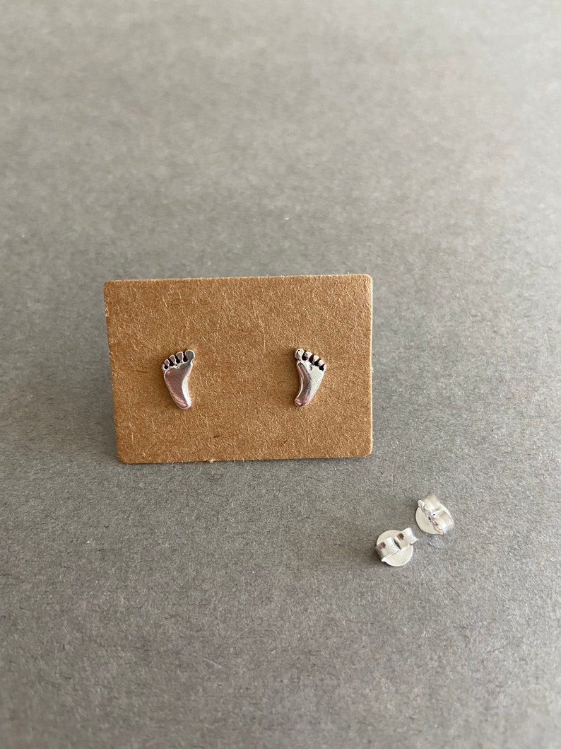 May include: A pair of silver stud earrings shaped like footprints. The earrings are displayed on a brown card.