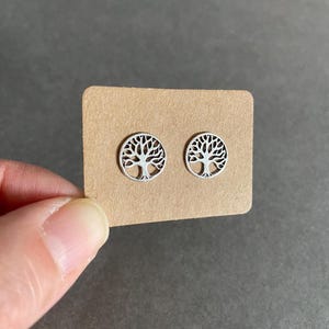 May include: A pair of silver stud earrings featuring a tree of life design. The earrings are displayed on a brown card.