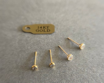 May include: Three gold stud earrings with small clear stones. The earrings are on a gray surface with a tag that reads "14KT GOLD".