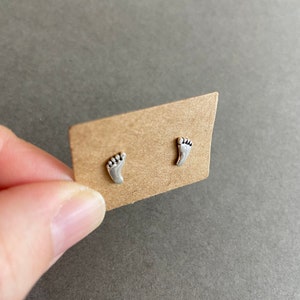 May include: A pair of silver stud earrings shaped like footprints. The earrings are displayed on a brown card.