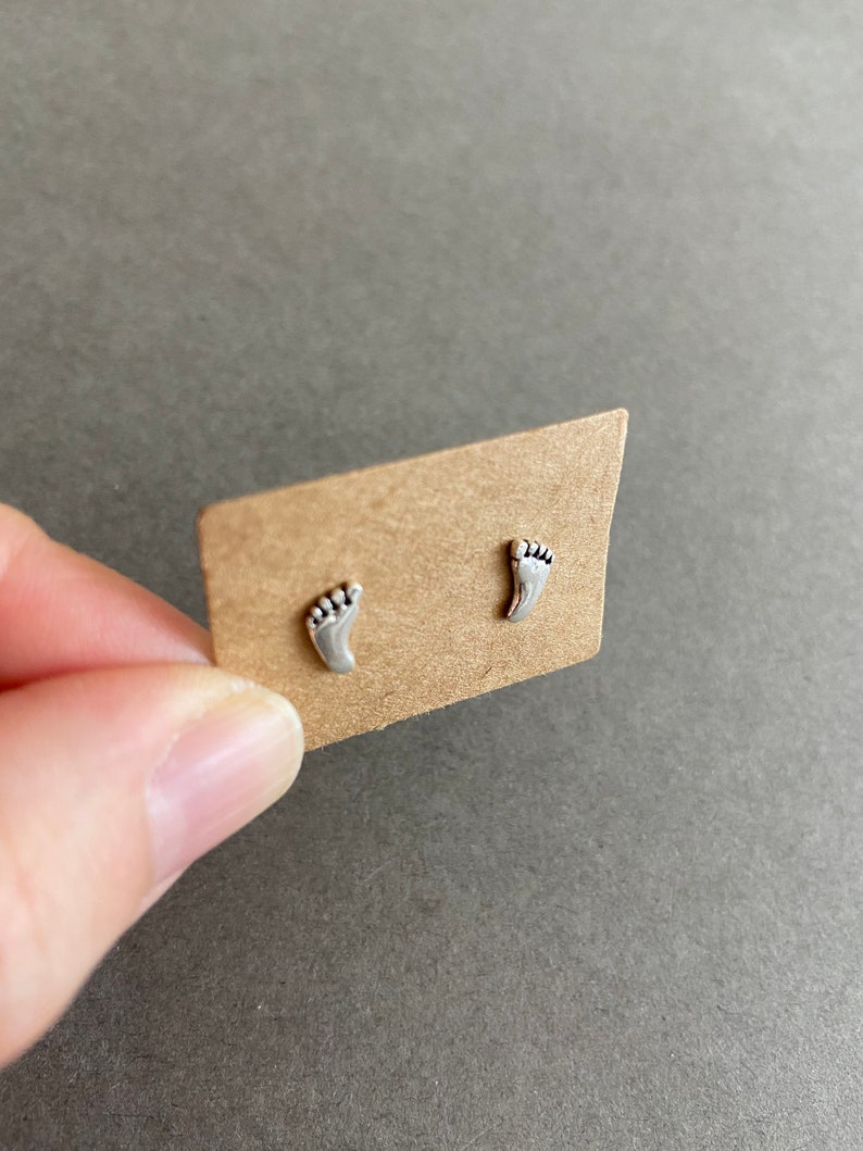 May include: A pair of silver stud earrings shaped like footprints. The earrings are displayed on a brown card.
