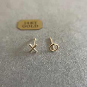May include: A pair of 14kt gold stud earrings featuring an X shape and an O shape. The earrings are positioned near a small rectangular tag with the text 14KT GOLD printed in dark letters. The studs have a polished finish and thin posts.