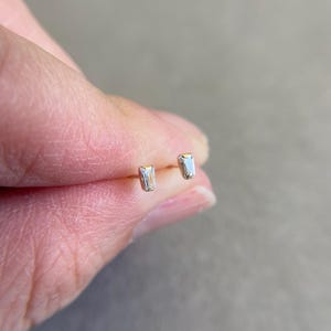 May include: A pair of gold stud earrings with a small, rectangular, clear gemstone in each earring.