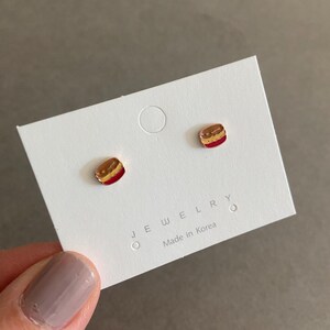 May include: A pair of gold-tone stud earrings with a red, brown, and yellow enamel design. The earrings are on a white card with the text "JEWELRY" and "Made in Korea".