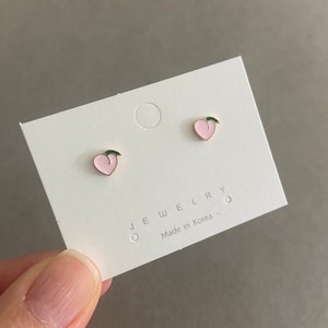 May include: A pair of peach-shaped earrings with gold accents and a green leaf detail. The earrings are on a white card with the text "JEWELRY Made in Korea".