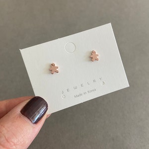 May include: A pair of gold-toned stud earrings featuring a small, minimalist person shape. The earrings are displayed on a white card with the text "JEWELRY" and "Made in Korea".