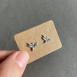 以下が含まれることがあります： 茶色のカードの上に置かれた、銀色のトンボのイヤリングのペア
