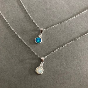 May include: Two silver necklaces with opal pendants. The top necklace has a blue opal pendant and the bottom necklace has a white opal pendant.