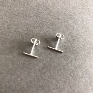 May include: A pair of silver stud earrings with a small horizontal bar. The earrings are simple and minimalist.