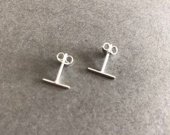 Può includere: Un paio di orecchini a lobo in argento con una piccola barra orizzontale. Gli orecchini sono semplici e minimalisti.