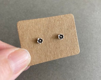 May include: A pair of small, silver-toned stud earrings shaped like five-petaled flowers with a central dot. The earrings are mounted on a rectangular piece of light brown, textured cardboard display card for presentation and storage.