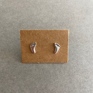May include: A pair of silver stud earrings featuring a footprint design. The earrings are displayed on a brown card.