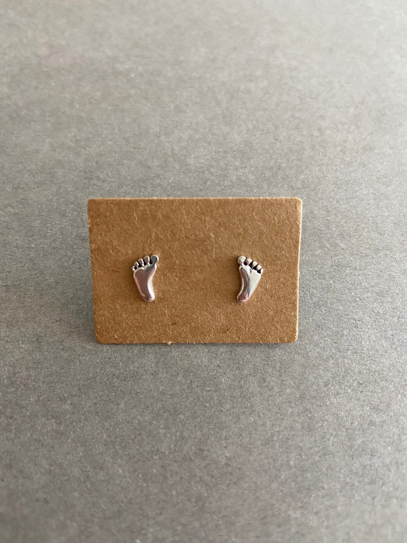 May include: A pair of silver stud earrings featuring a footprint design. The earrings are displayed on a brown card.