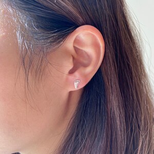 May include: A close-up of a person's ear with a small silver footprint earring. The earring is a simple design with a delicate outline.