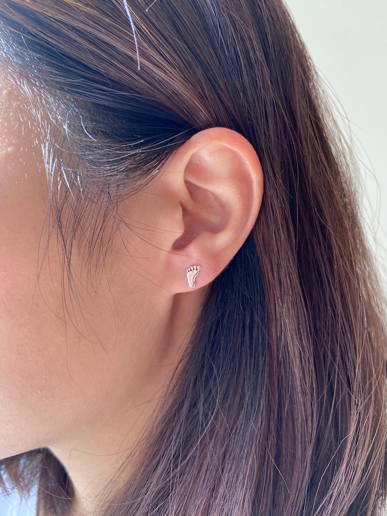 May include: A close-up of a person's ear with a small silver footprint earring. The earring is a simple design with a delicate outline.