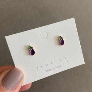 May include: A pair of small gold earrings with purple and green enamel eggplant designs. The earrings are on a white card with the text "JEWELRY" and "Made in Korea".