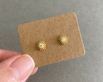 Peut inclure: Une paire de boucles d'oreilles clous en forme de soleil dorées avec un petit strass transparent au centre de chaque soleil.