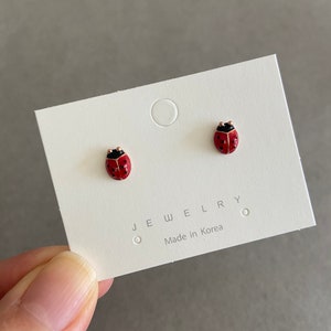 Può includere: Orecchini a bottone a forma di coccinella su un cartoncino bianco. Gli orecchini sono rossi con pois neri e teste nere. Il cartoncino recita "JEWELRY Made in Korea".