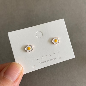 May include: A pair of gold-toned earrings shaped like fried eggs. The earrings have white yolks and yellow centres. The card reads "Jewelry" and "Made in Korea".