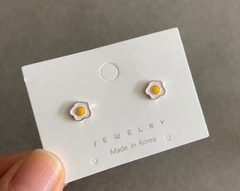 Könnte beinhalten: Ein Paar goldfarbene Ohrringe in Form von Spiegeleiern. Die Ohrringe haben weiße Eigelb und gelbe Zentren. Die Karte lautet "Schmuck" und "Hergestellt in Korea".