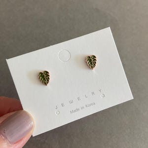 May include: Gold-toned leaf stud earrings with green enamel detail, displayed on a white card. The card reads "JEWELRY Made in Korea". The earrings are small, delicate, and feature a leaf design.