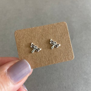 May include: A pair of small, silver-colored dragonfly stud earrings. The earrings are displayed on a brown cardboard backing, held by a hand with a purple painted fingernail. The dragonfly design features detailed wings and body.