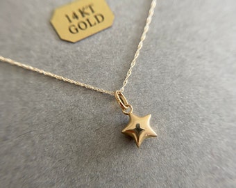May include: A gold-colored necklace with a star pendant. The star has a hollow center and is suspended from a delicate chain. A tag that reads "14KT GOLD" is visible in the background.
