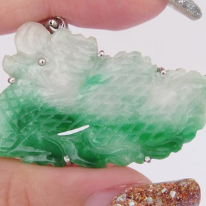 Pode incluir: Um pingente de jade verde e branco esculpido em forma de drag&atilde;o. O pingente &eacute; detalhado com esculturas intrincadas e tem uma argola de prata.