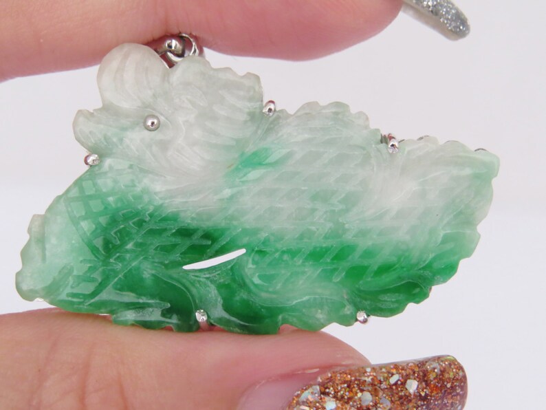 Pode incluir: Um pingente de jade verde e branco esculpido em forma de drag&atilde;o. O pingente &eacute; detalhado com esculturas intrincadas e tem uma argola de prata.