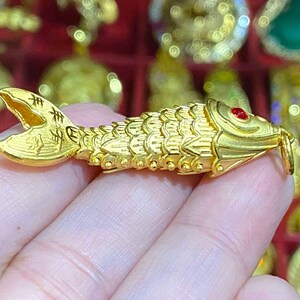 Puede incluir: Un amuleto de pez de color dorado con un ojo de piedra preciosa roja. El amuleto est&aacute; detallado con escamas y tiene caracteres chinos en la aleta de la cola.