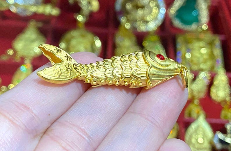 Puede incluir: Un amuleto de pez de color dorado con un ojo de piedra preciosa roja. El amuleto est&aacute; detallado con escamas y tiene caracteres chinos en la aleta de la cola.