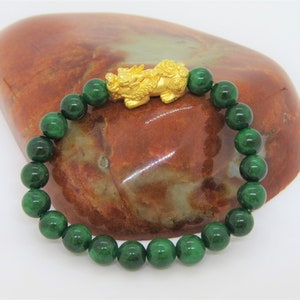Könnte beinhalten: Ein grünes Jade-Armband mit einem goldenen chinesischen Drachen-Anhänger. Das Armband liegt auf einem braunen Steinuntergrund.