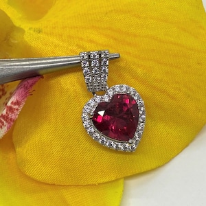 Könnte beinhalten: Ein herzförmiger Anhänger mit einem großen, tief roten Edelstein, der von einem Heiligenschein aus kleineren, klaren Edelsteinen umgeben ist. Der Anhänger ist aus Silber gefertigt und hat eine Öse zum Anbringen an einer Kette.