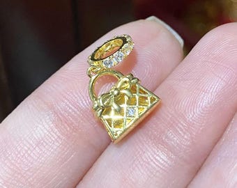 May include: Gold charm with a small purse design and a circle of clear stones. The purse has a diamond pattern and a bow on the top.